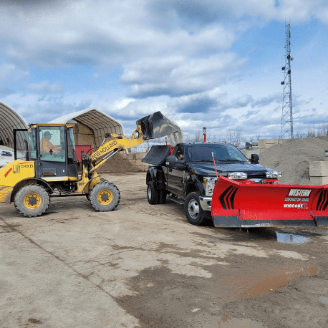 Yellow front-end loader filling a black pickup truck equipped with a large red snowplow.